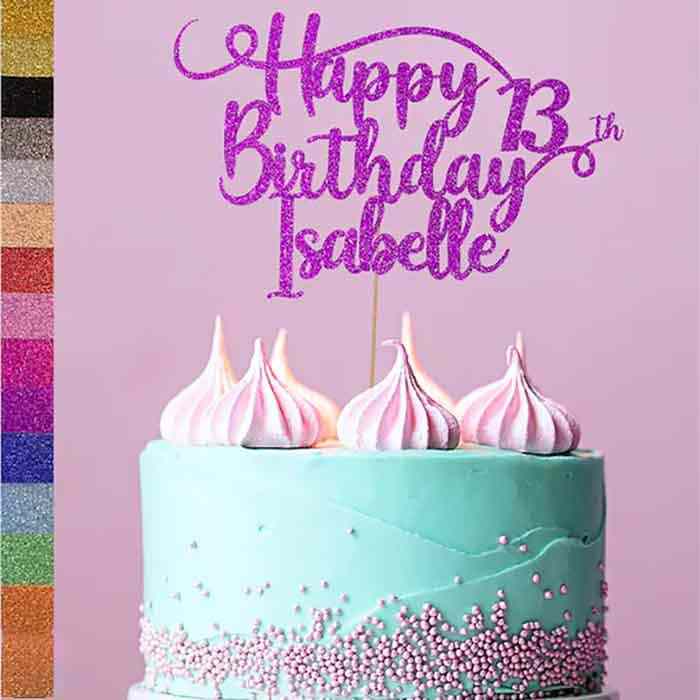 A birthday cake topped with a glittery cake topper that reads &quot;Happy Birthday Isabelle 13th&quot; in purple. The cake has a light blue frosting with pink and white meringue peaks on top, and is decorated with pink sugar pearls around the bottom. To the side, there are swatches of glitter in various colors. The background is a soft pink.