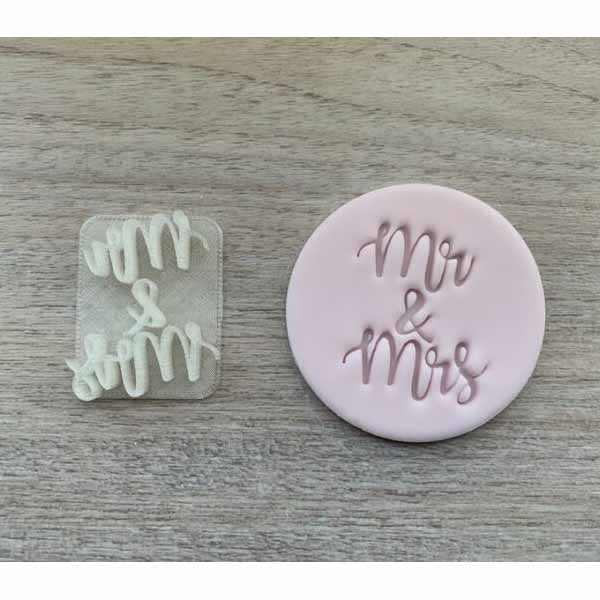 A pink fondant cookie stamp and a small clear plastic stamp. The pink stamp features the words &quot;Mr &amp; Mrs&quot; in a script font, and the clear stamp contains the same words &quot;&amp;&quot; in a smaller design. Both stamps are displayed on a light wooden surface.