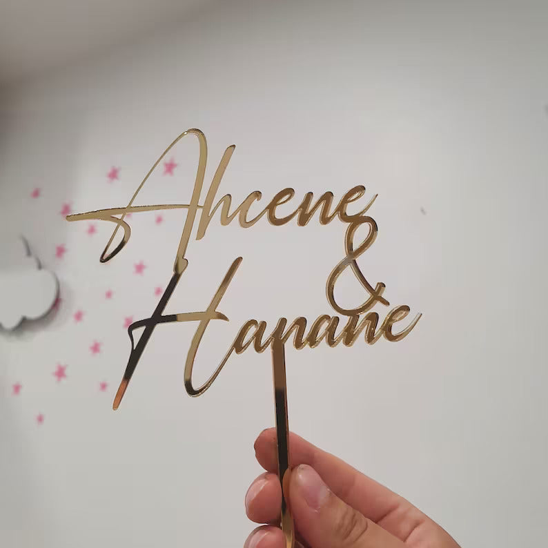 A hand is holding a personalized wedding cake topper made of gold-colored material. The topper features the names &quot;Ahcene&quot; and &quot;Hananë&quot; in elegant, cursive lettering, along with an ampersand symbol (&amp;) between the names. The background is light-colored with decorative elements like stars and clouds, enhancing the celebratory theme of the item.