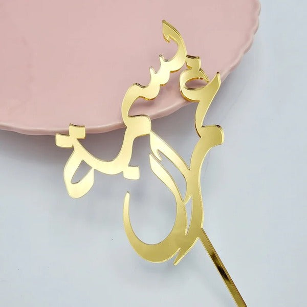 A gold acrylic cake topper featuring an elegant design, resting on a light pink plate. The topper has a stylized script that reads &quot;Happy Birthday&quot; in Arabic. The cake topper has a pointed stick for easy placement on cakes.