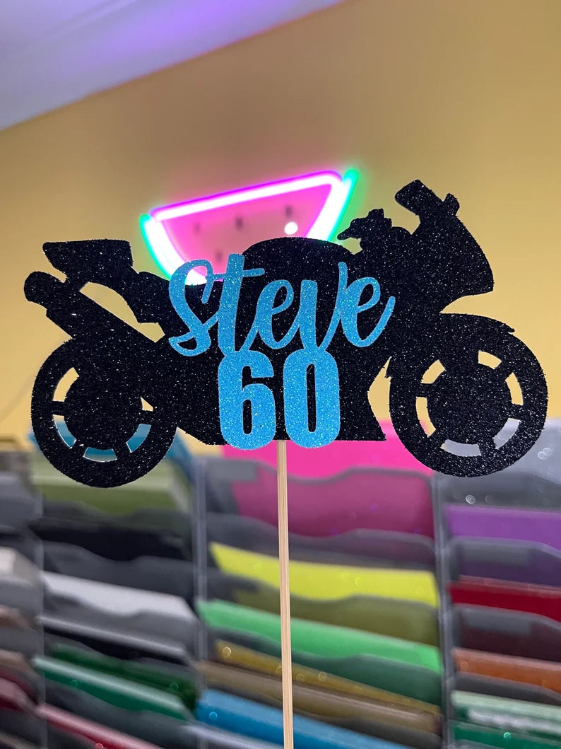 A cake topper made from black glitter cardstock features a motorcycle silhouette on either side. The text &quot;Steve&quot; is prominently displayed in a cursive blue font, with the number &quot;60&quot; in a bold blue font below it. The background shows colorful sheets of cardstock in various colors, and there is a soft glow of neon lights in the upper part of the image.