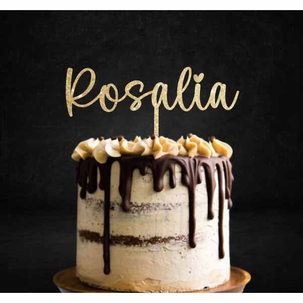 A decorated cake with a cream-colored frosting and chocolate ganache dripping down the sides. The cake is topped with a swirl of cream and banana slices. Above the cake, there is a sparkling gold glitter cake topper that reads &quot;Rosalia&quot; in a cursive font. The background is dark, enhancing the visual appeal of the cake.