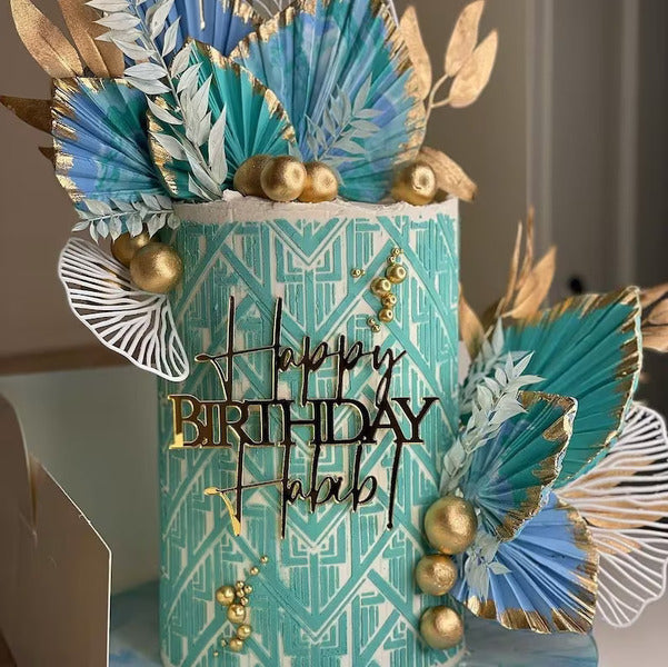 A beautifully decorated birthday cake charm featuring a cylindrical design in teal and gold colors. The cake is adorned with paper leaves in shades of blue and green, along with golden spheres. The phrase &quot;Happy Birthday&quot; is prominently displayed in elegant black lettering on the front, adding a festive touch. The overall design is vibrant and celebratory, perfect for a birthday celebration.