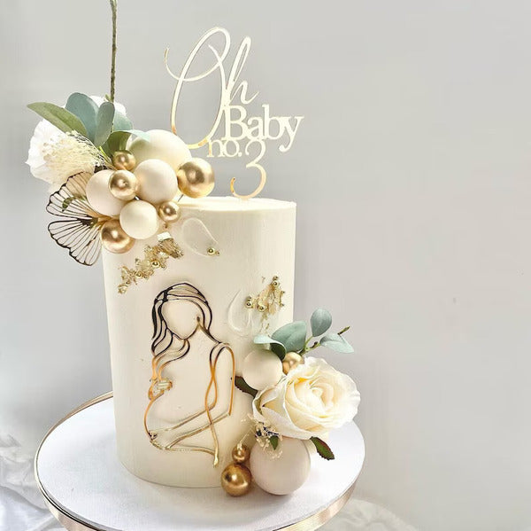 A decorated cake featuring a smooth white finish. At the top, there is a cake topper reading &quot;Oh Baby no. 3&quot; in an elegant font. Surrounding the cake are decorative elements including golden and white spherical accents, a white rose, greenery, and a delicate butterfly. The front of the cake features a minimalistic line drawing of a woman holding her stomach, symbolizing pregnancy. The overall aesthetic is soft and celebratory, suitable for a baby-themed event.