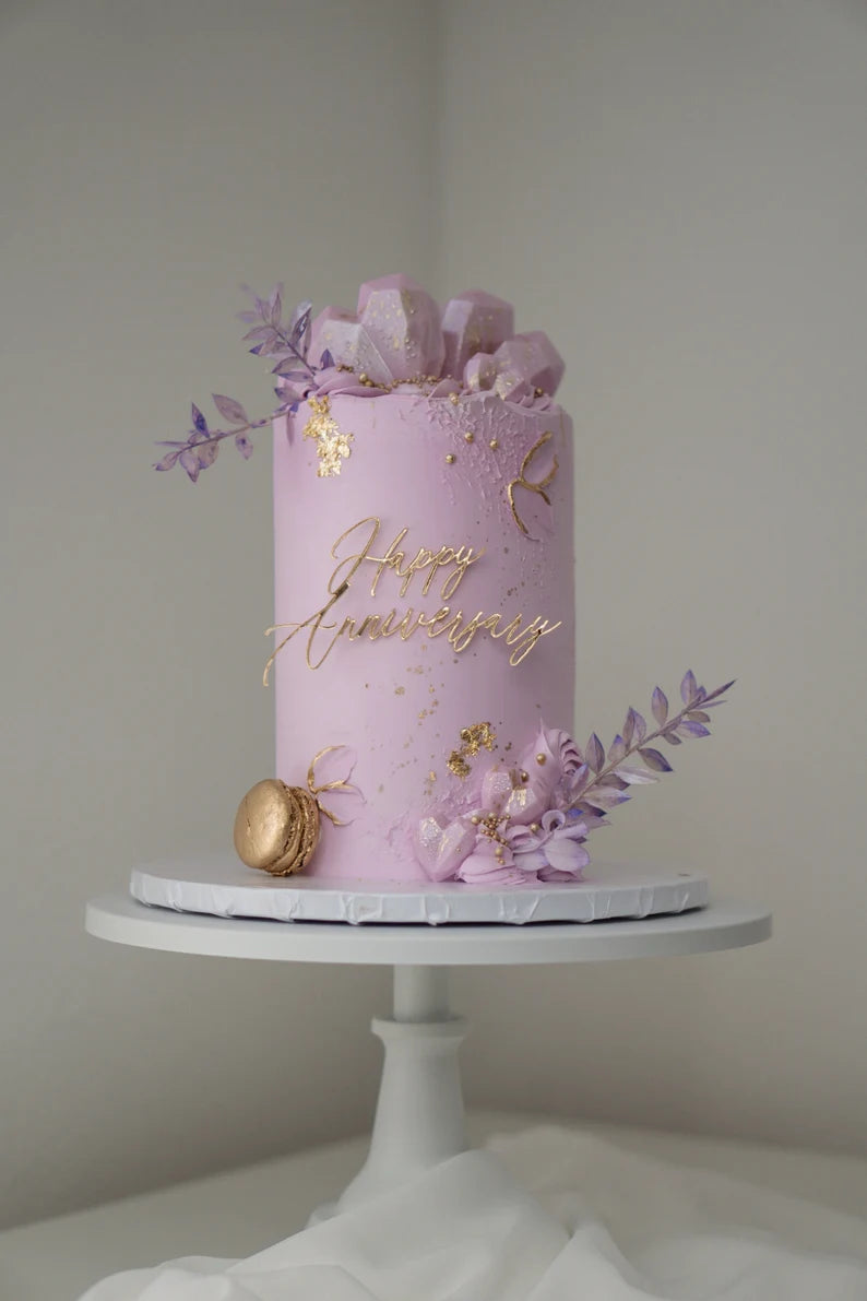 A beautifully decorated round cake sitting on a white cake stand. The cake is a soft lavender color and features a golden inscription that reads &quot;Happy Anniversary.&quot; Adorning the top of the cake are edible decorations, including geometric candy pieces, purple and lavender flowers, and gold accents. A gold macaron is also placed at the base of the cake, completing the elegant design. The background is neutral, emphasizing the cake&