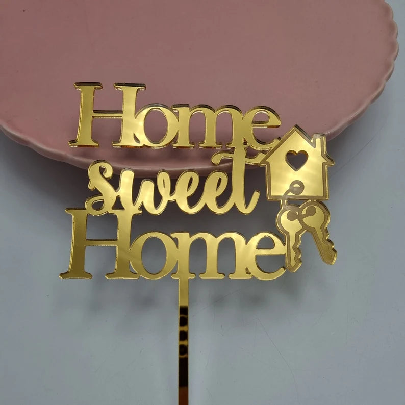 A golden acrylic cake topper featuring the text &quot;Home sweet Home.&quot; The letters are designed with a playful font, and there is a small house icon with a heart and two keys attached to the design. The topper is placed near the edge of a pink plate.