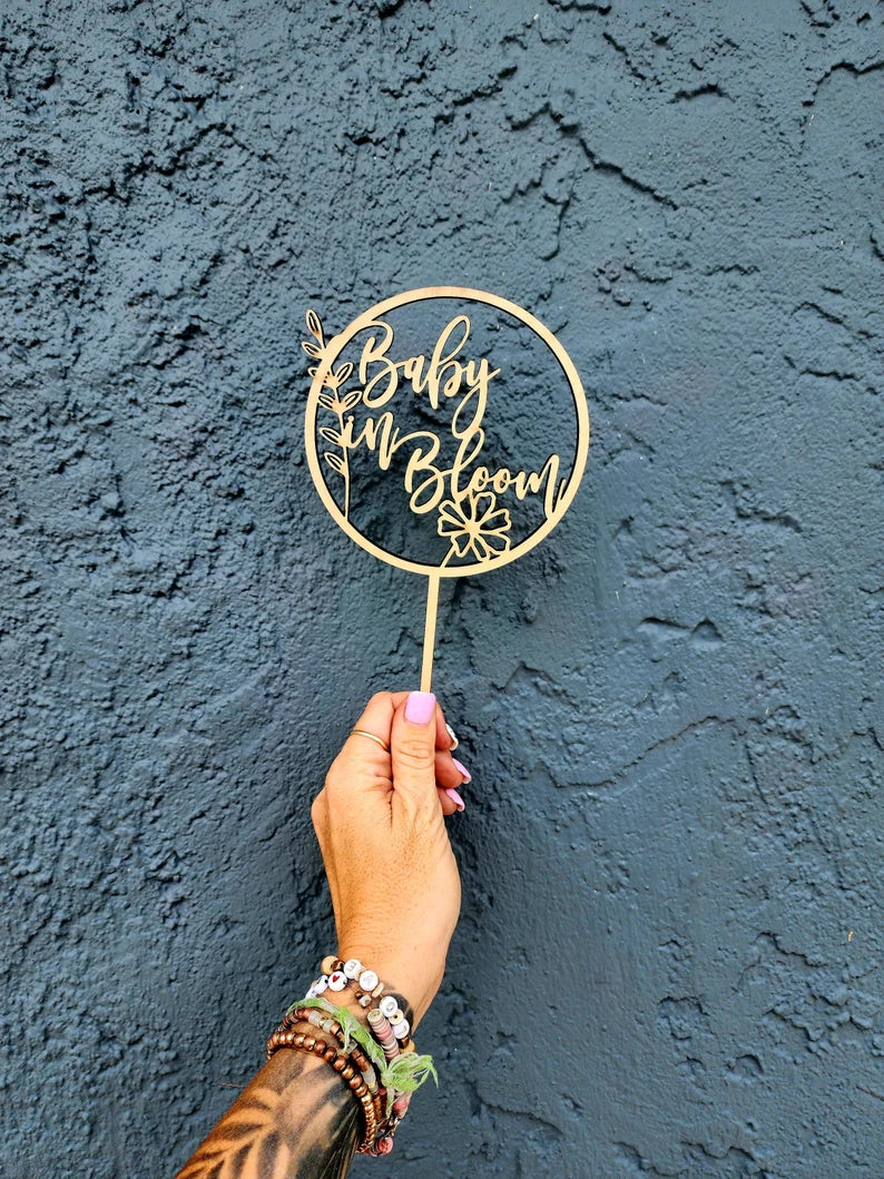 A hand is holding an acrylic cake topper that features the words &quot;Baby in Bloom&quot; inside a circular design. The topper includes decorative elements, such as flowers and leaves. The hand is adorned with multiple colorful bracelets, and the background is a textured dark blue wall.