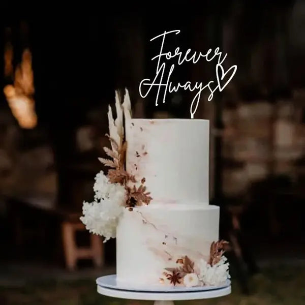 A two-tiered, white wedding cake is displayed on a round pedestal. The cake features decorative elements, including sprigs of dried foliage and clusters of small white flowers on one side. Atop the cake, a stylish cake topper reads &quot;Forever Always&quot; in elegant, cursive script, accompanied by a small heart symbol. The background is softly blurred, suggesting an outdoor setting.