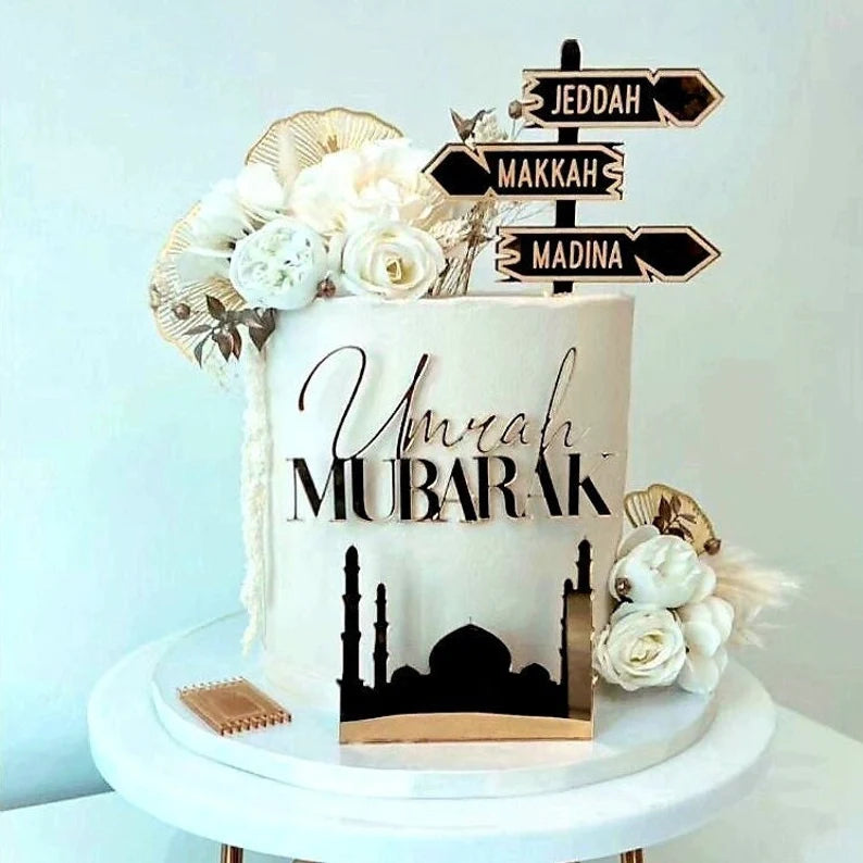 The image features a decorative cake with a cream-colored exterior. At the front, the words &quot;Umrah Mubarak&quot; are beautifully crafted in an elegant font. The cake is adorned with white and pale floral arrangements along with golden accents. Topping the cake, there is a signpost with three arrows indicating the directions to &quot;Jeddah,&quot; &quot;Makkah,&quot; and &quot;Madina.&quot; Additionally, a silhouette of a mosque is depicted at the base of the cake, enhancing its festive theme. The cake is displayed on a white round stand.