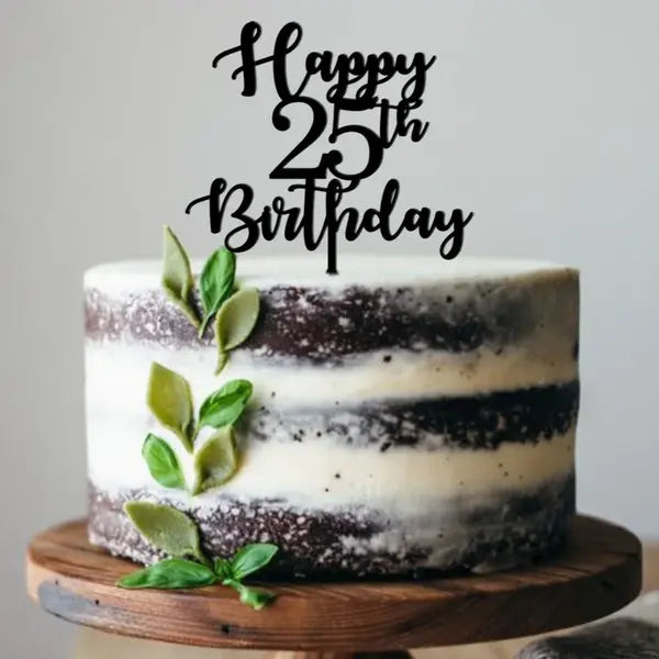 A tiered cake with a light cream and dark chocolate exterior, topped with a black acrylic cake topper that reads &quot;Happy 25th Birthday.&quot; The cake is decorated with green leaves along the side and sits on a round wooden cake stand. The background is softly blurred.