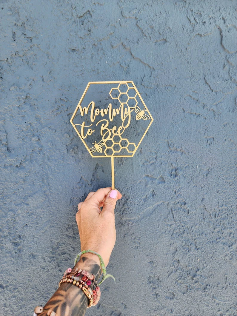 An acrylic cake topper featuring the words &quot;Mommy to Bee&quot; in a stylish font, surrounded by honeycomb patterns and small bee illustrations. The topper is golden in color and held up against a textured blue wall. A hand adorned with multiple bracelets is holding the topper from its stick base.