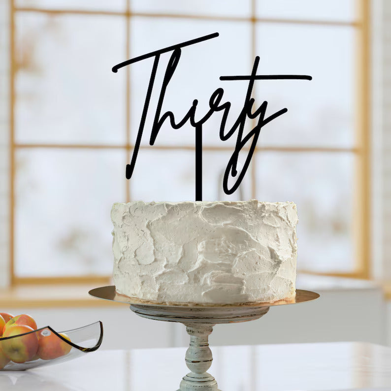 A round cake with a textured white frosting sits on a decorative pedestal stand. Atop the cake is a black acrylic topper that reads &quot;Thirty&quot; in an elegant cursive font. The background features a window with soft lighting, and a plate with apples is visible in the foreground.