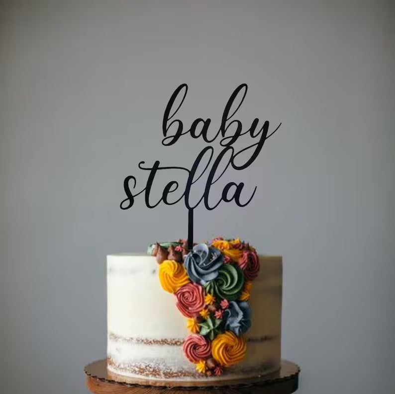 A two-tiered cake with a smooth white frosting and a partially exposed cake layer is topped with a black acrylic cake topper that reads ?baby stella? in an elegant script. The cake is decorated on one side with colorful buttercream roses and small flowers in shades of pink, yellow, and green. The cake sits on a wooden round cake stand against a neutral background.