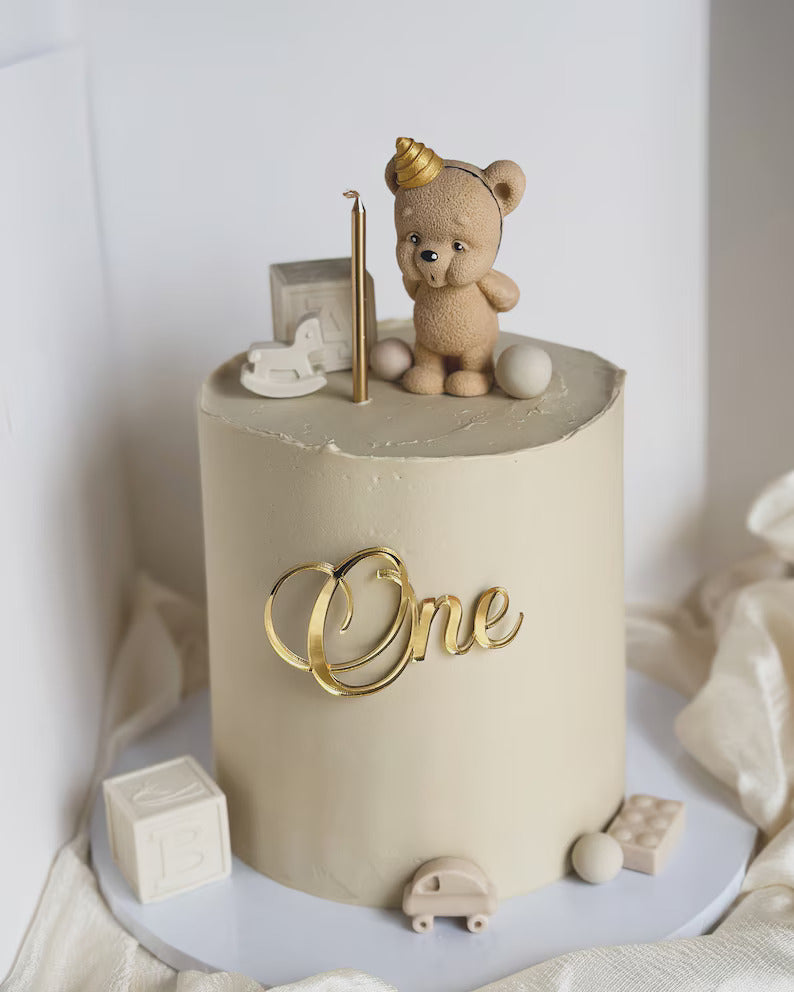 A minimalist, cream-colored cake is decorated on top with a small, tan bear figurine wearing a golden cone hat. The bear stands next to a gold-colored candle, with a small toy rocking horse and round decorative balls nearby. The cake&