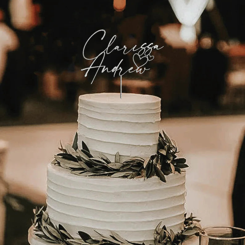 A three-tiered wedding cake with a smooth white icing is adorned with a decorative trim of greenery around its base. Atop the cake is a personalized acrylic cake topper featuring the names &quot;Clarissa&quot; and &quot;Andrew,&quot; elegantly written in script, along with two simple heart shapes. The background suggests a festive event setting with soft lighting.