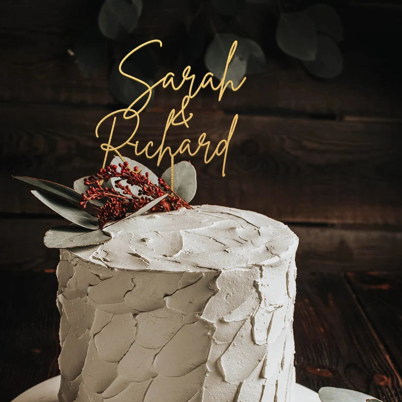 A round wedding cake with a textured gray frosting, topped with a golden acrylic cake topper that reads &quot;Sarah &amp; Richard.&quot; The cake is decorated with eucalyptus leaves and small red berries, set against a dark wooden background.