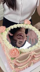 The image shows a beautifully decorated cake with a mirror-like surface in the center, surrounded by decorative icing flowers in soft pink and cream colors. A person is positioned above the cake, reflected in the mirror surface, appearing to smile and gesturing with their hands. The cake is presented on a flat surface, and the overall setting looks festive and celebratory.