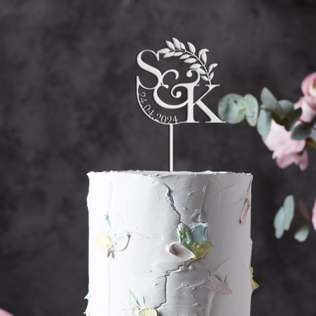 A close-up of a white, textured wedding cake featuring pastel-colored flower decorations. Atop the cake is a custom acrylic cake topper displayed in silver, featuring the initials "S&amp;K" and the date "24.04.2024," accompanied by a delicate leaf design. The background is softly blurred, enhancing the focus on the cake and topper.