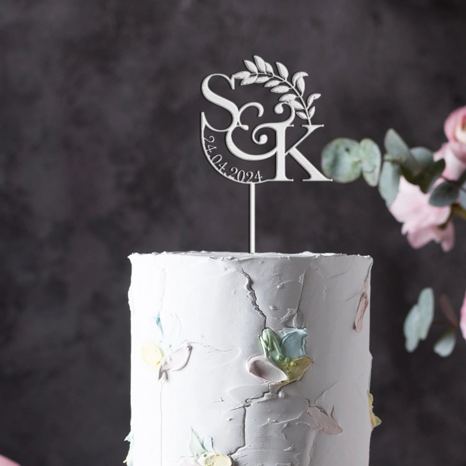 A close-up of a white, textured wedding cake featuring pastel-colored flower decorations. Atop the cake is a custom acrylic cake topper displayed in silver, featuring the initials "S&amp;K" and the date "24.04.2024," accompanied by a delicate leaf design. The background is softly blurred, enhancing the focus on the cake and topper.