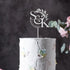 A close-up of a white, textured wedding cake featuring pastel-colored flower decorations. Atop the cake is a custom acrylic cake topper displayed in silver, featuring the initials "S&K" and the date "24.04.2024," accompanied by a delicate leaf design. The background is softly blurred, enhancing the focus on the cake and topper.