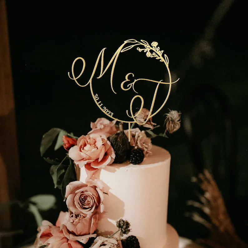 An elegant wedding cake adorned with a personalized acrylic topper featuring the initials "M&amp;D" and the date "30.11.2023." The cake is decorated with soft pink and purple roses, dark berries, and greenery, creating a romantic and floral aesthetic. The background is dark, highlighting the beauty of the cake and the topper.