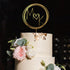 A three-tiered white wedding cake topped with a gold acrylic cake topper featuring the initials "M" and "K" inside a circular design with a heart in between. The cake is adorned with delicate pink and white flowers, as well as eucalyptus leaves, and has gold leaf accents on the top tier. The background is dark, highlighting the cake and its decorations.