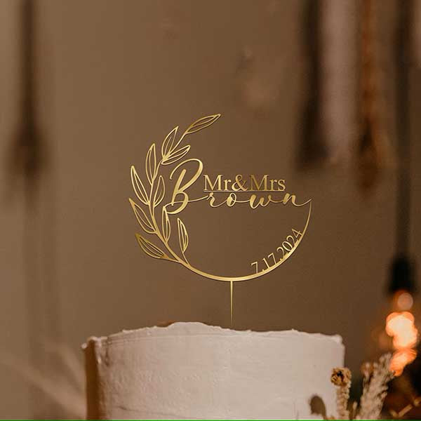 A golden acrylic cake topper featuring the text &quot;Mr &amp; Mrs Brown&quot; with decorative leaves and the date &quot;7.17.2024.&quot; The topper is placed on a white wedding cake in a softly lit setting.