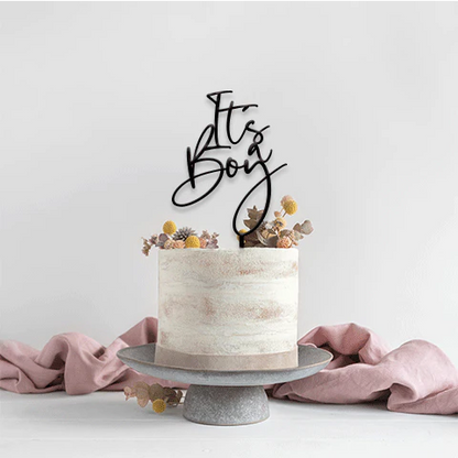 A round, two-tiered cake with a textured cream-colored frosting is decorated with dried flowers on top. A black acrylic cake topper that says "It&