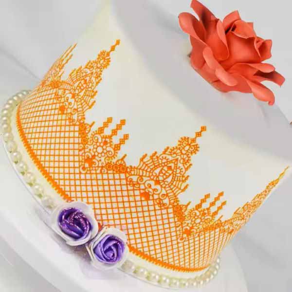 A decorated cake with a smooth white surface and an intricate embossed design in orange along the bottom edge. The design features a floral lace pattern resembling a crown. At the top, there is a large orange flower, and at the base, two small roses in purple and white. The cake is set on a round white cake stand adorned with a string of small pearls.