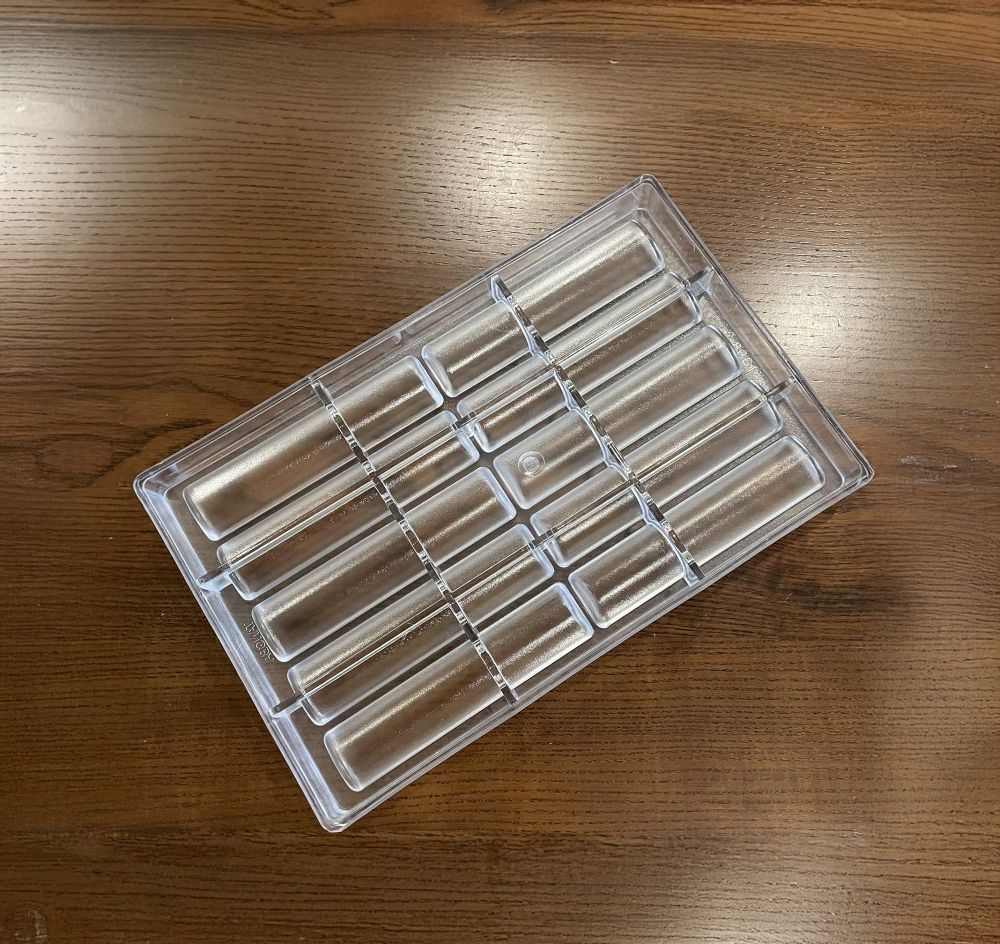 Clear plastic ice mold tray with multiple rectangular slots sits on a wooden table. The light wood grain adds a warm contrast to the transparent tray.