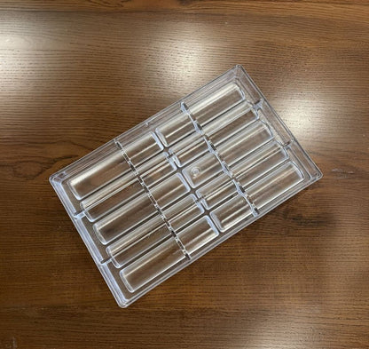 Clear plastic ice mold tray with multiple rectangular slots sits on a wooden table. The light wood grain adds a warm contrast to the transparent tray.