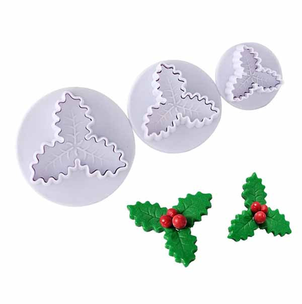 A set of three plunger cutters designed to create holly leaf shapes, displayed alongside three finished holly decorations made from green fondant with red berries. The cutters are white with leaf designs in the center, showing varying sizes from large to small, while the decorations exhibit vibrant green leaves and bright red berries.