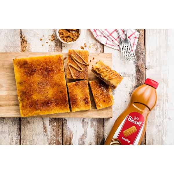 An image of a wooden cutting board displaying a baked dessert cut into squares, topped with a caramelized surface. There are several pieces of the dessert arranged on the board, with swirls of topping drizzled on top. A bottle of Lotus Biscoff topping is prominently placed on the right side. In the background, there is a small bowl containing crushed biscuit crumbs, a checkered napkin, and a pair of forks, all set on a rustic wooden table.