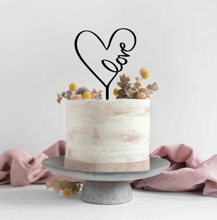 A round, two-tiered cake with a textured white frosting is displayed on a gray cake stand. Atop the cake is a black acrylic topper in the shape of a heart with the word &quot;love&quot; written inside it. The cake is decorated with small dried flowers and foliage, featuring shades of yellow, pink, and green, adding a delicate touch. A soft pink fabric is draped on the surface beside the cake, enhancing the overall aesthetic.