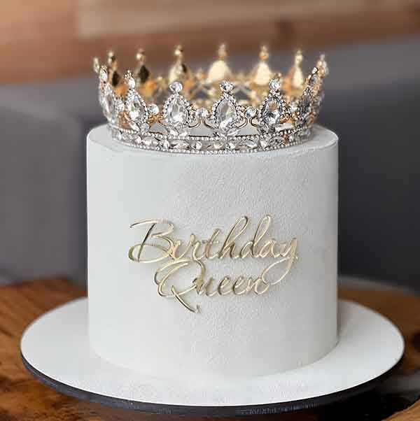 A round, white birthday cake is topped with a sparkling silver and gold crown. The cake features the elegant text "Birthday Queen" in a cursive, golden font, prominently displayed on its front. The background is softly blurred, emphasizing the cake and crown.