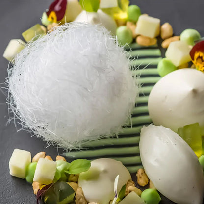 A delicate dessert presented on a plate with a white spun sugar ball, creamy dollops, diced fruits, nuts, and green striped garnish, creating an elegant, gourmet display.