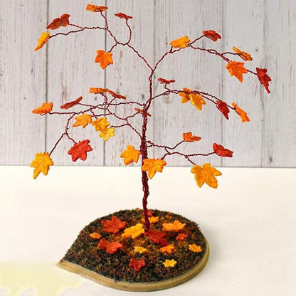 Image description for accessibility: A decorative miniature tree with branches made of thin wire, adorned with small maple leaves in various autumn colors, including red, orange, and yellow. The tree is positioned on a circular base covered with brown material resembling soil, also scattered with additional small leaves in similar colors. The background features a light wooden panel.