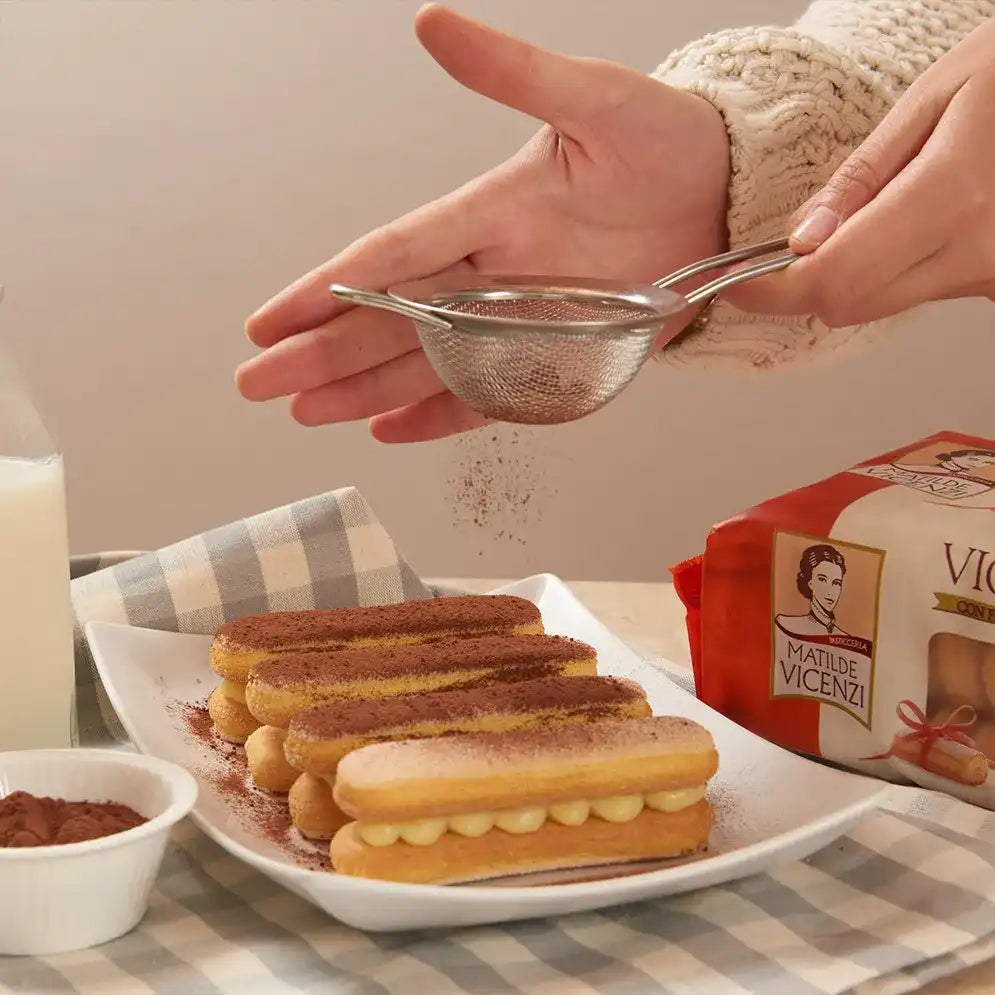 Hand sprinkling cocoa powder over a plate of Savoiardo Vicenzovo Ladyfingers filled with cream, alongside a pack of Matilde Vicenzi Ladyfingers, a milk bottle, and a bowl of cocoa powder on a checkered cloth.