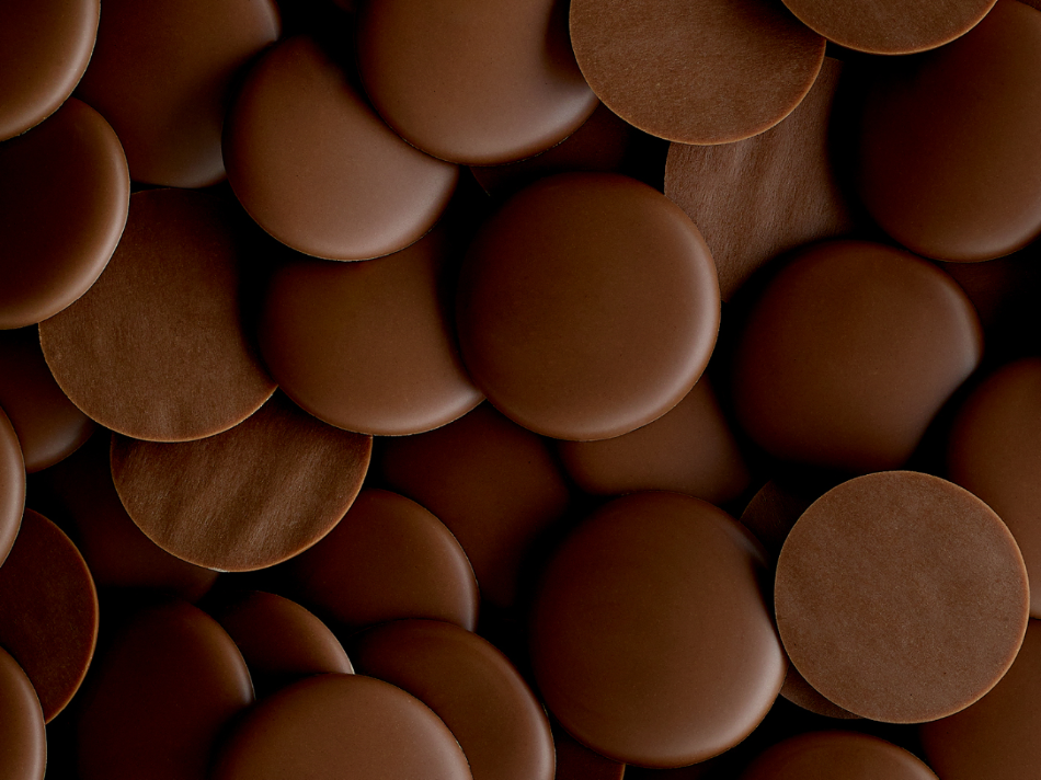 A close-up image of many round milk chocolate discs, varying in size and slightly overlapping. The discs have a smooth texture and a rich, dark brown color, creating a visually appealing pattern. The overall look is enticing and emphasizes the chocolate&