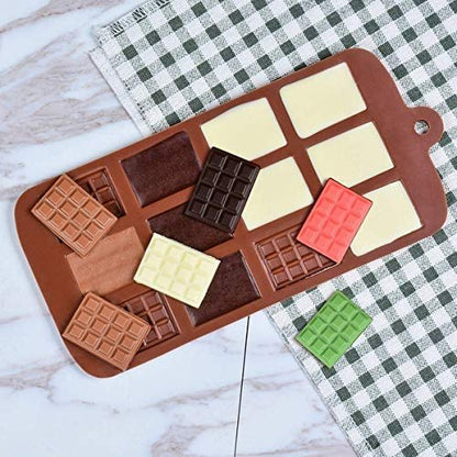 A silicone chocolate bar mold featuring various mini chocolate shapes in brown, red, green, and cream colors. Some chocolate pieces are placed both inside the mold and around it, with a checkered green and white cloth visible in the background. The mold has a tab for easy handling.