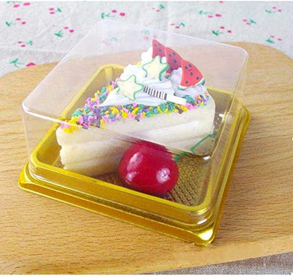 A small dessert displayed in a clear plastic box. The dessert resembles a slice of cake, topped with colorful sprinkles, white icing, and small decorative elements including green stars and red shapes that appear to be strawberries. Below the cake slice, there is a shiny red cherry placed on a textured golden base. The box is set on a wooden surface with a light floral fabric underneath.