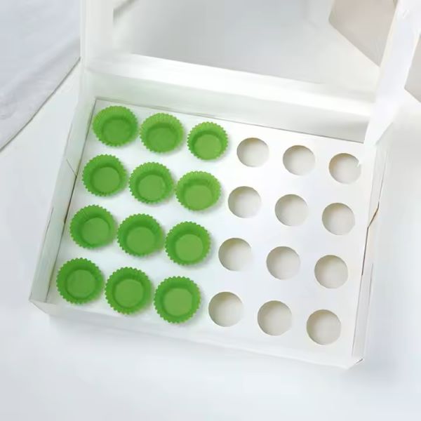 The image shows a white cupcake box designed to hold 24 mini cupcakes. Inside the box, there are 12 green silicone cupcake liners arranged in two rows on the left side. The right side of the box has space for 12 additional cupcakes, indicated by empty circular holes. The background is neutral and light, emphasizing the box and its contents.