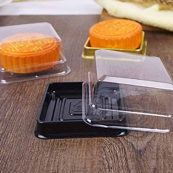 A display of transparent plastic boxes used for packaging mooncakes. There are two mooncakes visible?one orange and one yellow, both with decorative patterns on top. The boxes include a black base box and a clear cover, positioned in various orientations on a wooden surface.