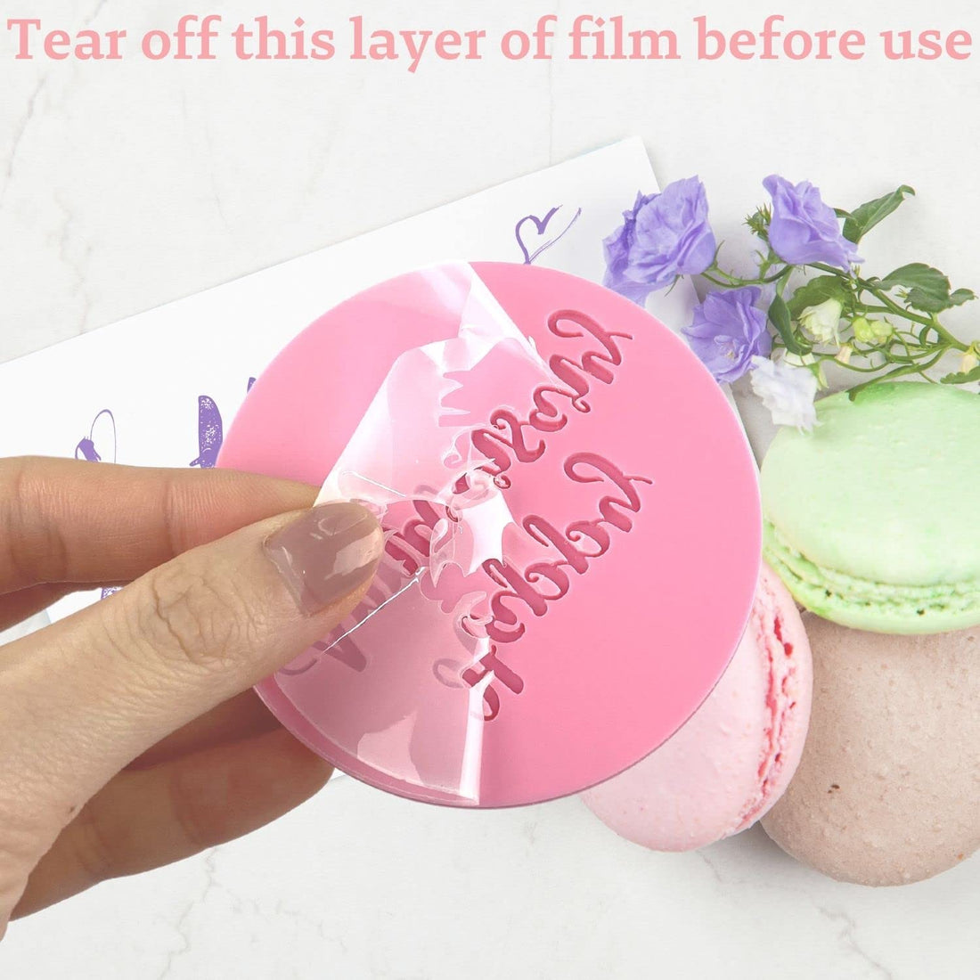 A hand is holding a round pink fondant embosser cookie stamp with the words "Mr &amp; Mrs" embossed on it. A transparent protective film is being partially peeled off the top of the stamp. In the background, there are colorful macarons (pink, green, and brown) and small purple flowers. At the top of the image, there is a message in light pink text that reads "Tear off this layer of film before use."