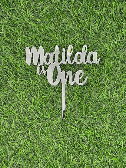 A decorative acrylic cake topper featuring the text "Matilda is One" in a stylish script font. The topper has a stake for easy insertion into a cake. It is displayed against a background that resembles green grass or artificial turf.