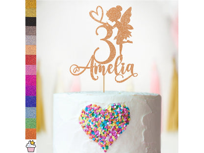 A decorative cake topper featuring a glittery fairy figure beside the number "3" and the name "Amelia," displayed on a white frosted cake. The cake is adorned with colorful heart-shaped sprinkles in shades of pink, blue, and yellow. To the left, a color palette of glitter cardstock options is shown in various vibrant hues, including gold, silver, black, and several shades of pink and purple. The background features soft, blurred decorations.