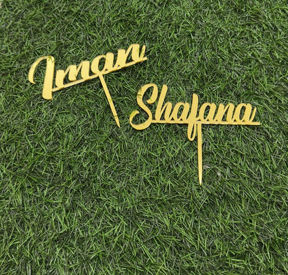 Two gold cake toppers with the names "Imran" and "Shafana" displayed on a green grass-like background. The toppers feature an elegant, cursive font with pointed ends for easy insertion into a cake.