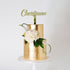 A two-tiered cake with a shiny gold finish, topped with a customized name topper reading "Christiaan." The cake is adorned with white flowers and green foliage, creating an elegant presentation on a white cake stand against a plain white background.