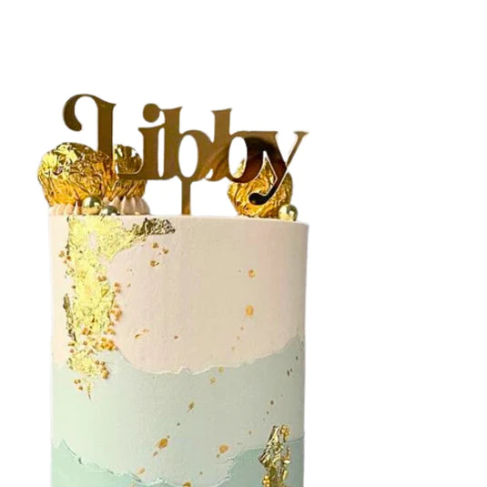 A round cake decorated with layers of pale pink and light blue frosting, adorned with gold foil accents. At the top, a golden acrylic cake topper displays the name &quot;Libby&quot; in an elegant script font. The cake is also topped with decorative gold-wrapped candies.