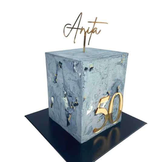 A gray marble-patterned cake in the shape of a cube, prominently displaying the number &quot;50&quot; in gold on its front face. Atop the cake is a customized acrylic topper with the name &quot;Anita&quot; in a cursive gold font. The cake is placed on a black square base.